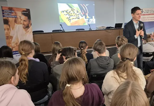 Grupa młodych osób siedzi w sali konferencyjnej, zwrócona przodem do mężczyzny z mikrofonem prowadzącego prezentację. Przed nimi ekran z wyświetlonym obrazem i stolik z mikrofonami. Po lewej stronie widoczny stojak z plakatem.