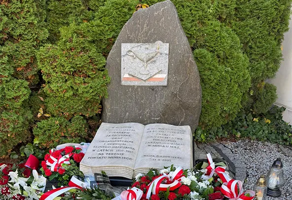 Pomnik z dużym kamieniem i otwartą rzeźbioną księgą na podstawie, otoczony zielonymi krzewami. Wokół kamienia i księgi leżą wiązanki kwiatów z biało-czerwonymi wstążkami oraz kilka zniczy.