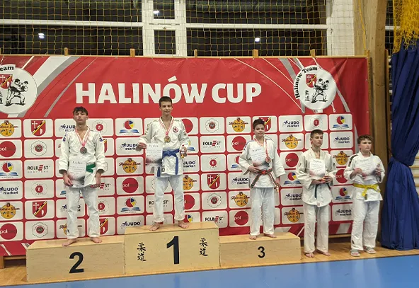 Pięciu chłopców w białych strojach do judo stoi na podium i obok niego. Trzech na podium z medalami na szyi, pozostali dwaj trzymają dyplomy. Tło czerwone z napisem HALINÓW CUP i logotypami. Wnętrze hali sportowej.