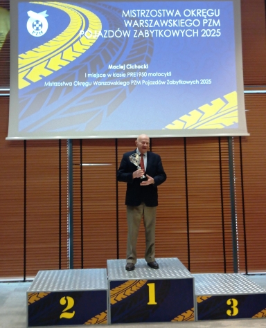 Mężczyzna stoi na podium na miejscu pierwszym i trzyma puchar. Nad nim wisi duży niebiesko-żółty ekran z motywem bieżnika opony. W tle pionowe brązowe żaluzje i widoczne podium z polami oznaczonymi cyframi 1, 2 i 3.