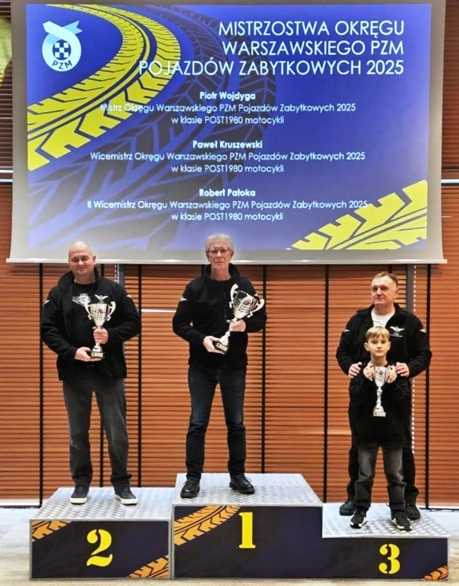 Trzech mężczyzn i chłopiec stoją na podium z numerami 1, 2 i 3, każdy trzyma puchar. W tle jest duży niebieski baner z grafiką bieżnika opon; scena odbywa się wewnątrz, na jasnym tle drewnianych paneli.