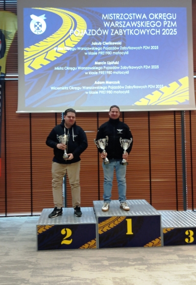 Dwóch mężczyzn stoi na podium wewnątrz sali, na pierwszym i drugim stopniu. Obaj trzymają puchary, za nimi wisi duży ekran z kolorową grafiką, a tło stanowią zamknięte drewniane żaluzje.