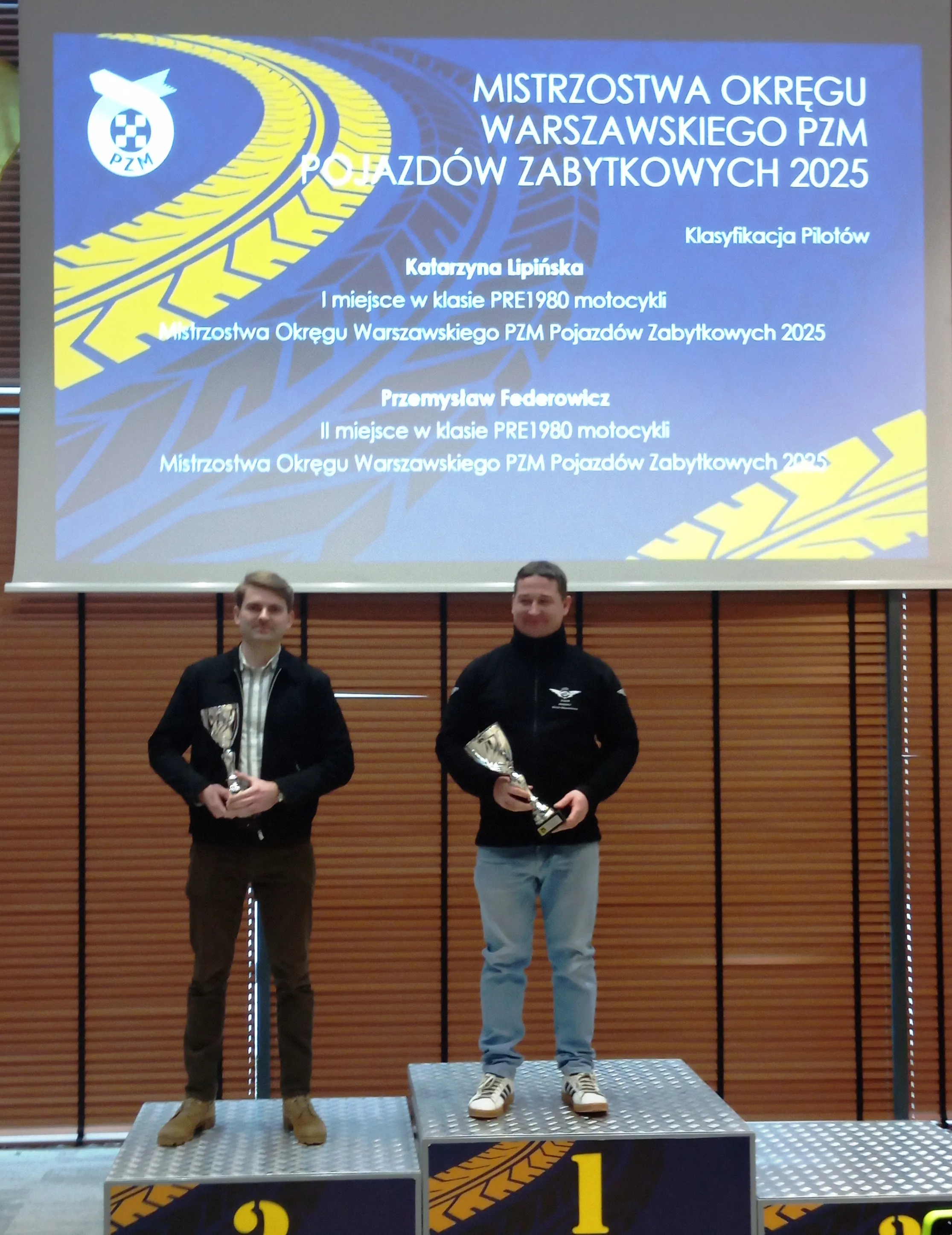 Dwóch mężczyzn stoi na podium w hali. Mężczyzna po prawej stoi na pierwszym stopniu i trzyma puchar. Mężczyzna po lewej stoi na drugim stopniu i trzyma puchar. Nad nimi wisi duży niebieski ekran z żółym wzorem opony i białym tekstem. W tle drewniane żaluzje.