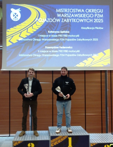 Dwóch mężczyzn stoi na podium w hali. Mężczyzna po prawej stoi na pierwszym stopniu i trzyma puchar. Mężczyzna po lewej stoi na drugim stopniu i trzyma puchar. Nad nimi wisi duży niebieski ekran z żółym wzorem opony i białym tekstem. W tle drewniane żaluzje.