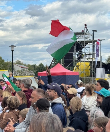Tłum ludzi na zewnątrz podczas wydarzenia, z osobą machającą flagą w kolorach czerwonym, białym i zielonym, pod pochmurnym niebem.