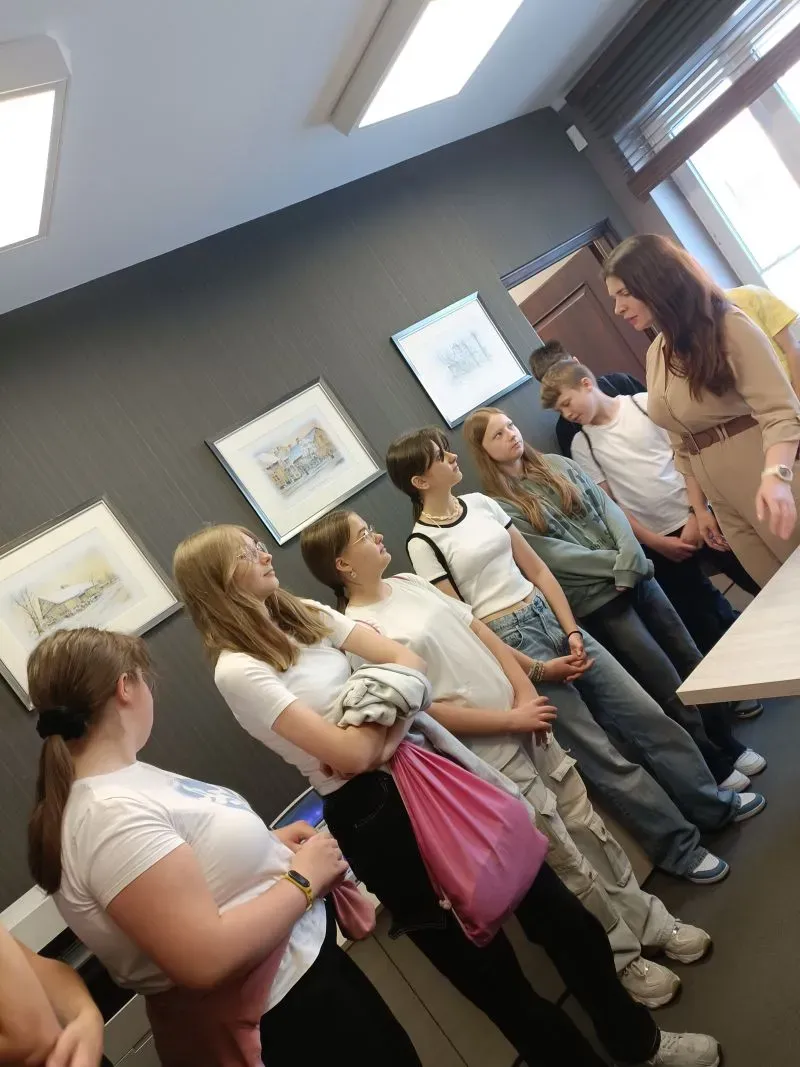 Grupa młodych osób stoi w pomieszczeniu i rozmawia z kobietą w beżowej sukience.