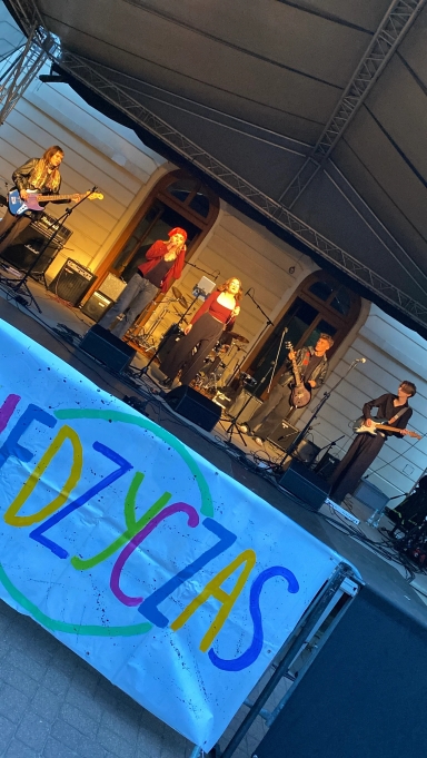 Piątka muzyków na scenie plenerowej pod namiotem, grających na instrumentach i śpiewających, na pierwszym planie kolorowy baner z napisem.