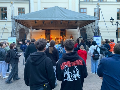 Grupa ludzi stoi na zewnątrz przed sceną z zadaszeniem, na której występuje zespół muzyczny. W tle budynek z dużymi oknami. Wieczorne światło. Publiczność ubrana głównie w ciemne kurtki i bluzy.