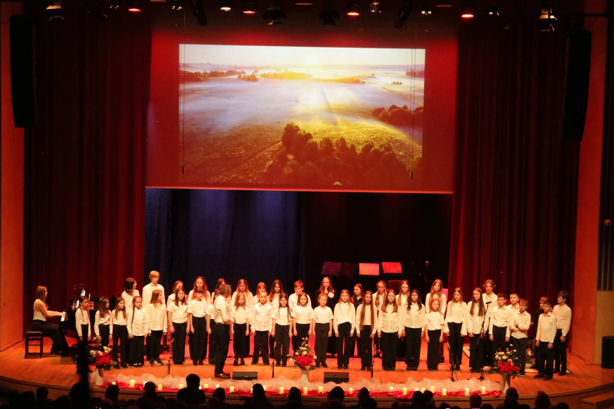 Grupa dzieci i nastolatków stoi na scenie teatralnej w rzędzie, ubrani na biało i czarno. Z lewej strony gra osoba przy fortepianie. W tle na dużym ekranie wyświetlany jest obraz krajobrazu z widokiem na poranne pola i drzewa oświetlone wschodzącym słońcem. Na pierwszym planie stoją kwiaty i świece. Publikum siedzi na widowni. Scena jest oświetlona ciepłym, czerwonym światłem.