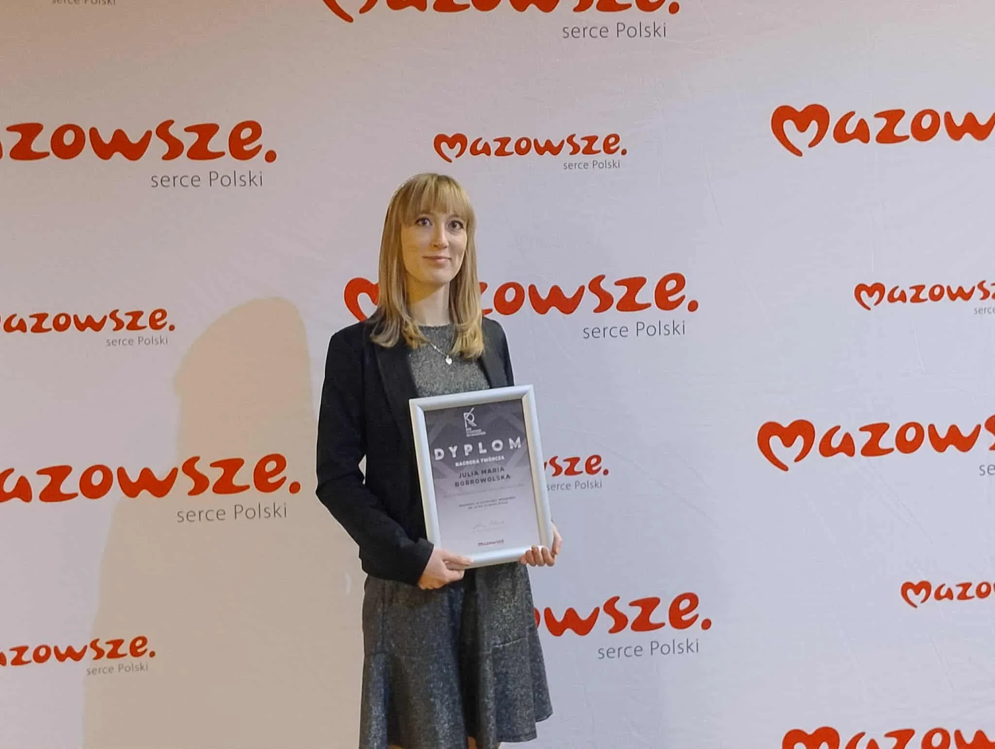 Kobieta stoi na tle białego bannera z napisem „Mazowsze serce Polski” w kolorze czerwonym, trzyma w dłoniach dyplom w srebrnej ramie, ubrana w szarą sukienkę i czarną marynarkę.