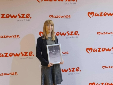 Kobieta stoi na tle białego bannera z napisem „Mazowsze serce Polski” w kolorze czerwonym, trzyma w dłoniach dyplom w srebrnej ramie, ubrana w szarą sukienkę i czarną marynarkę.