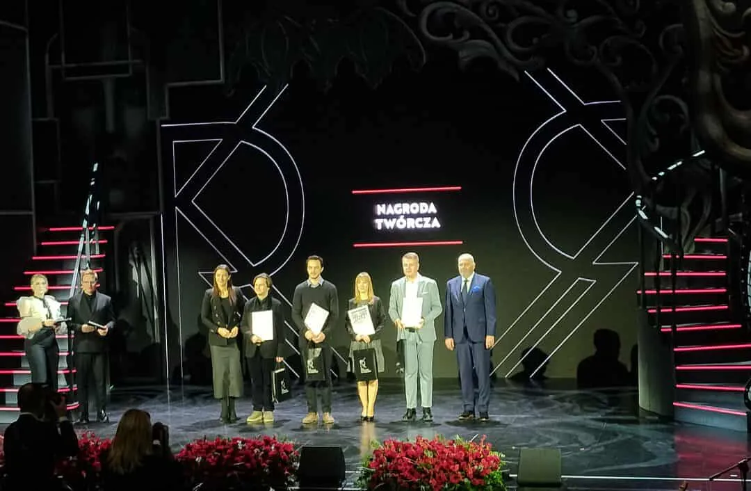 Siedem osób stoi na scenie przy ciemnym tle z jasnymi liniami, za nimi napis "Nagroda Twórcza". Na pierwszym planie czerwona dekoracja kwiatowa. Po bokach sceny schody z czerwonymi podświetleniami. Widownia częściowo widoczna na dole.