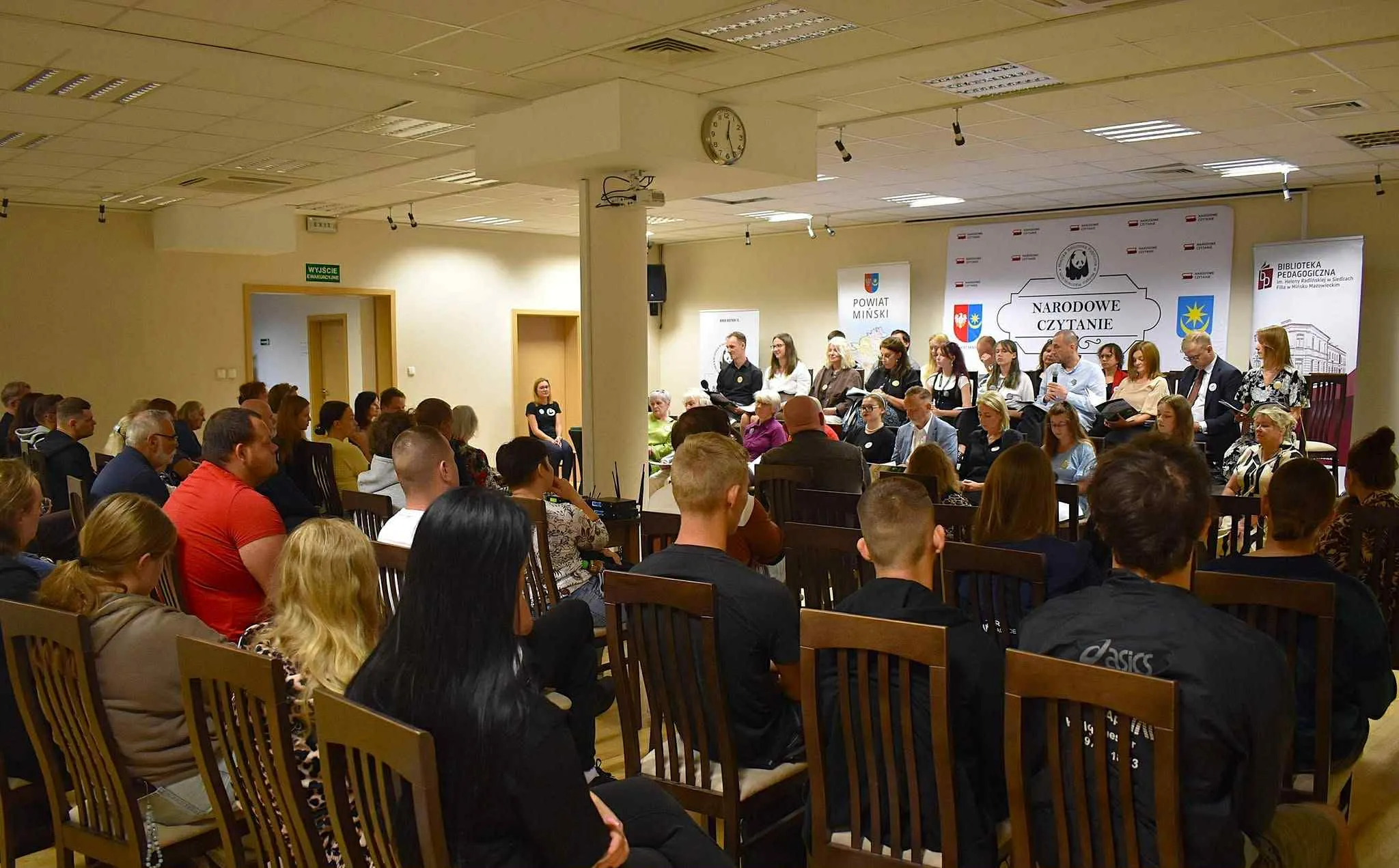 Grupa osób siedzi na krzesłach w sali podczas wydarzenia Narodowe Czytanie, część z nich czyta teksty na scenie z mikrofonem.