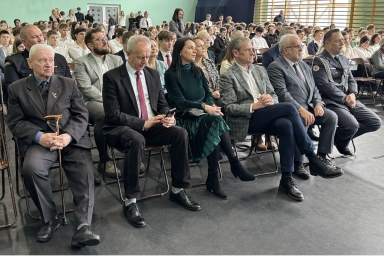 Grupa osób siedzi w rzędach krzeseł na sali gimnastycznej. W pierwszym rzędzie siedzi starszy mężczyzna z laską, mężczyzna w garniturze z telefonem, kobieta w zielonej sukience oraz trzej mężczyźni w marynarkach i mundurze. W tle siedzi wiele młodych osób w białych koszulach przy zielonej ścianie.