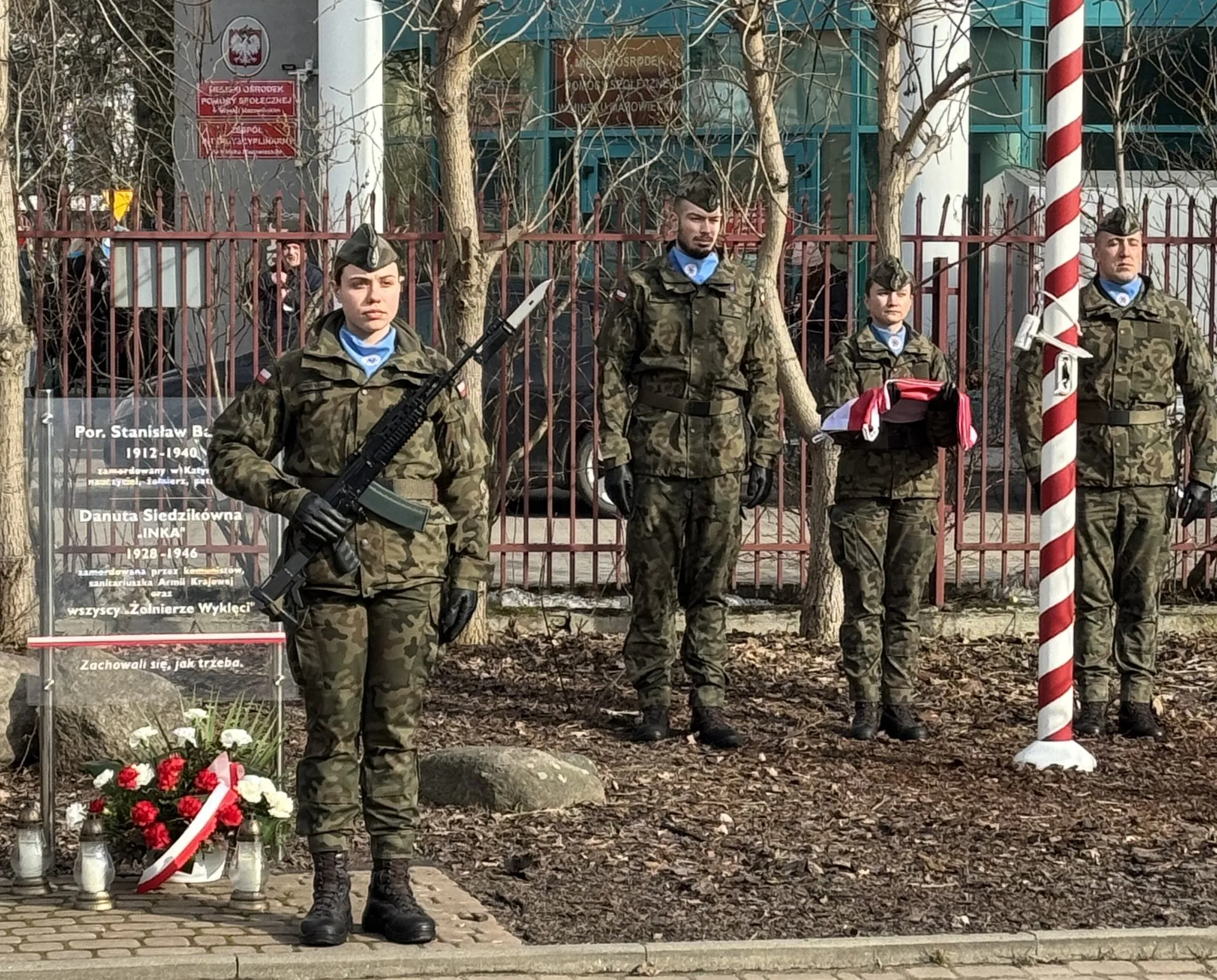 Czterech umundurowanych żołnierzy stoi na zewnątrz w rzędzie przed metalowym ogrodzeniem. Po lewej żołnierka trzyma karabin z bagnetem. Druga od prawej trzyma złożoną biało-czerwoną flagę. Po lewej przy chodniku leży wieniec z czerwono-białymi kwiatami i zapalonymi zniczami. Po prawej stoi pionowy maszt w biało-czerwone paski. W tle nagie drzewa i budynek.
