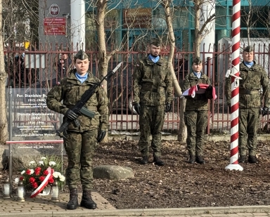 Czterech umundurowanych żołnierzy stoi na zewnątrz w rzędzie przed metalowym ogrodzeniem. Po lewej żołnierka trzyma karabin z bagnetem. Druga od prawej trzyma złożoną biało-czerwoną flagę. Po lewej przy chodniku leży wieniec z czerwono-białymi kwiatami i zapalonymi zniczami. Po prawej stoi pionowy maszt w biało-czerwone paski. W tle nagie drzewa i budynek.