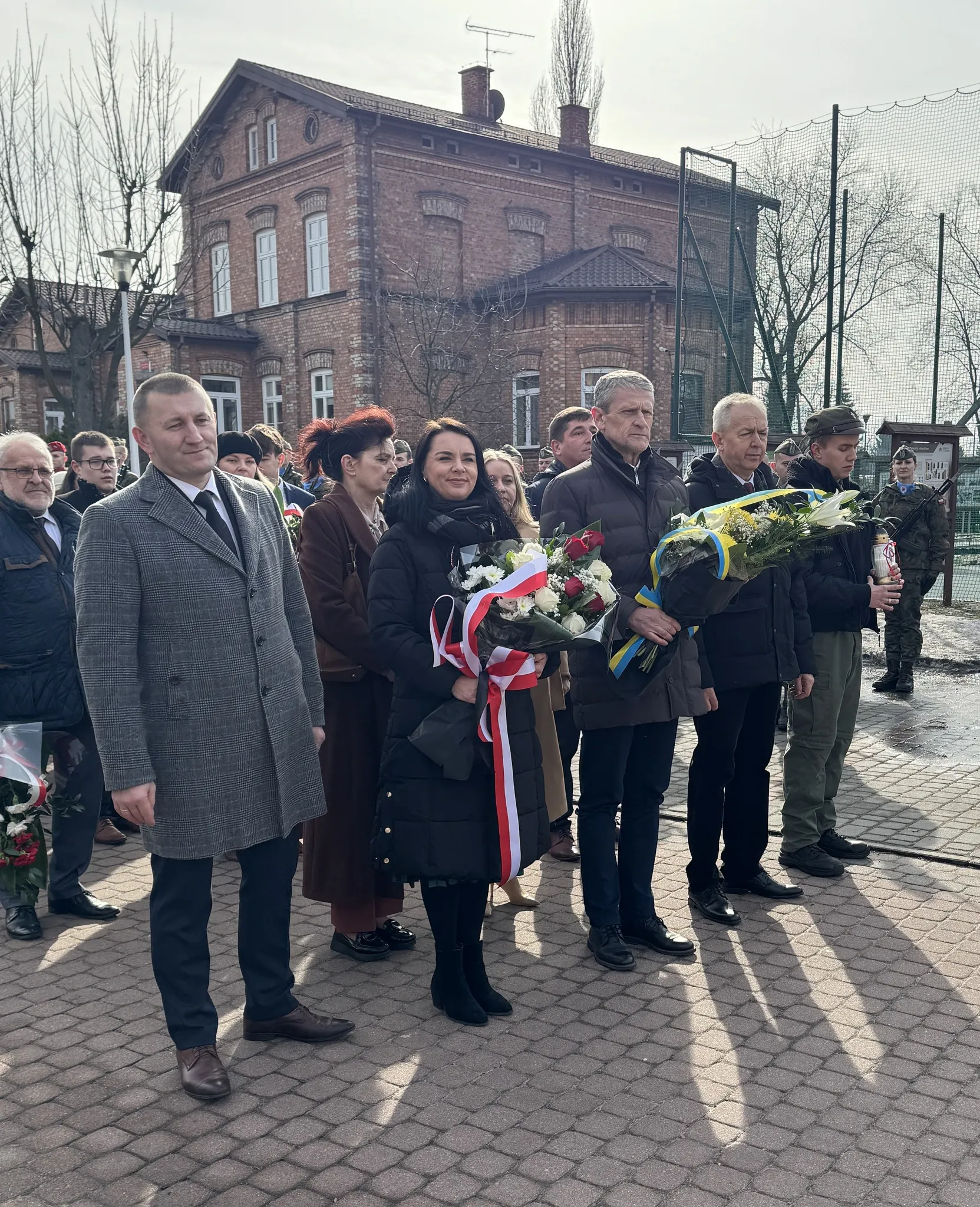 Grupa osób stoi na zewnątrz przed ceglanym budynkiem w ciągu dnia. W pierwszym rzędzie pięć dorosłych osób trzyma wiązanki kwiatów z biało‑czerwonymi i niebiesko‑żółtymi wstążkami. W tle kilkoro obserwatorów i żołnierz przy ogrodzeniu. Niebo jest pochmurne.