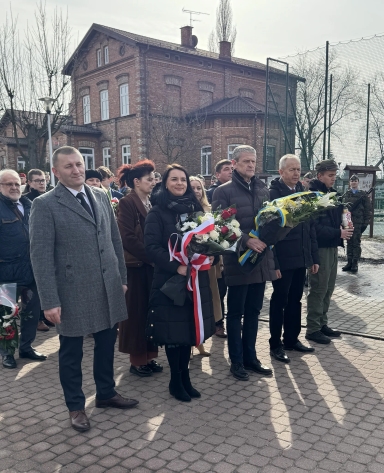 Grupa osób stoi na zewnątrz przed ceglanym budynkiem w ciągu dnia. W pierwszym rzędzie pięć dorosłych osób trzyma wiązanki kwiatów z biało‑czerwonymi i niebiesko‑żółtymi wstążkami. W tle kilkoro obserwatorów i żołnierz przy ogrodzeniu. Niebo jest pochmurne.