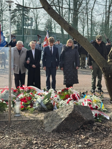 Grupa pięciu osób stoi przy ziemi z ułożonymi wieńcami i zniczami. Po prawej stoi mężczyzna w wojskowym mundurze, w tle widoczne flagi i kilka umundurowanych osób. Przed nimi na ziemi leży duży kamień i gałąź drzewa przebiega skośnie przez obraz.