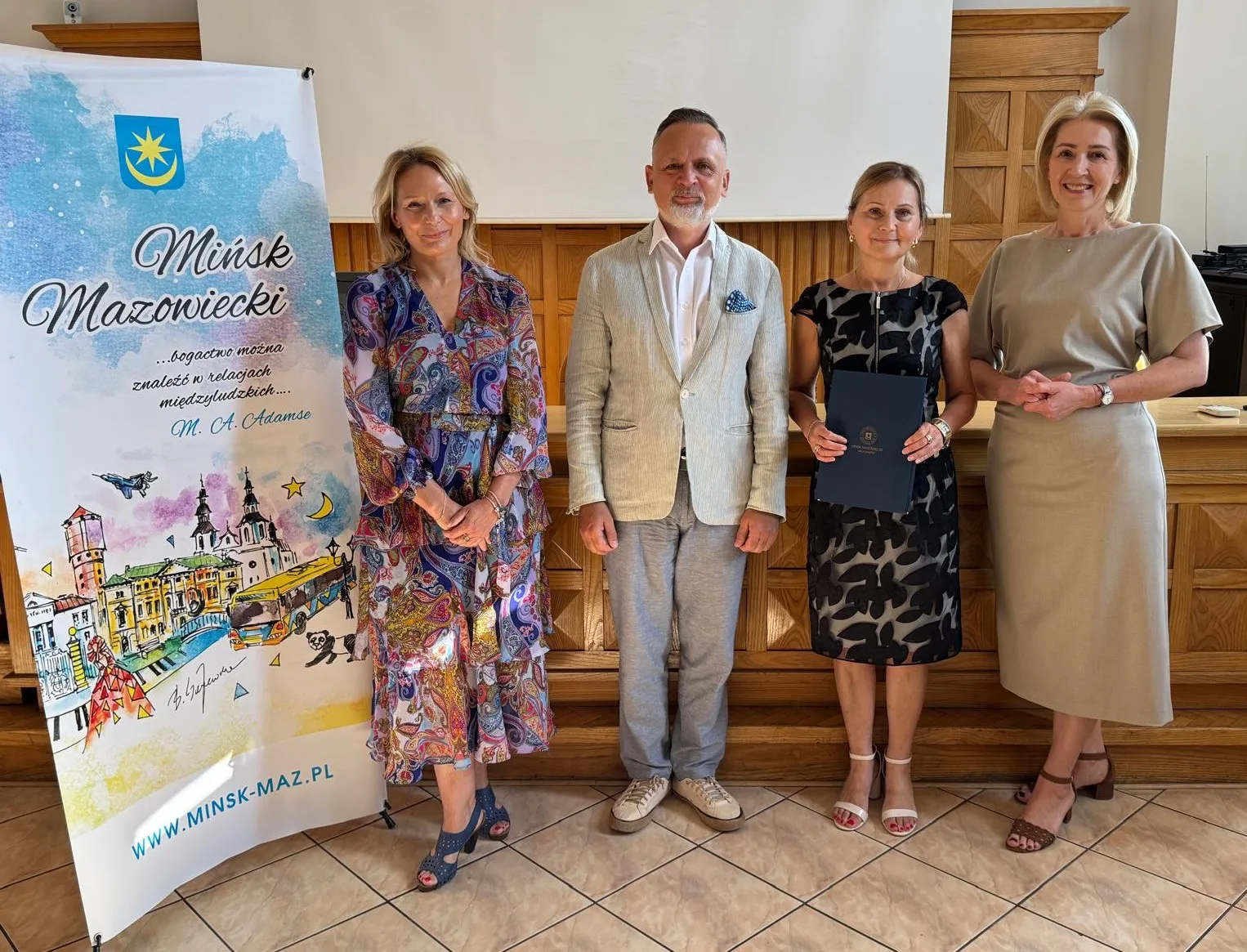 Cztery osoby stoją w pomieszczeniu przy drewnianej ścianie obok stojaka z planszą o Mińsku Mazowieckim, jedna kobieta trzyma teczkę.