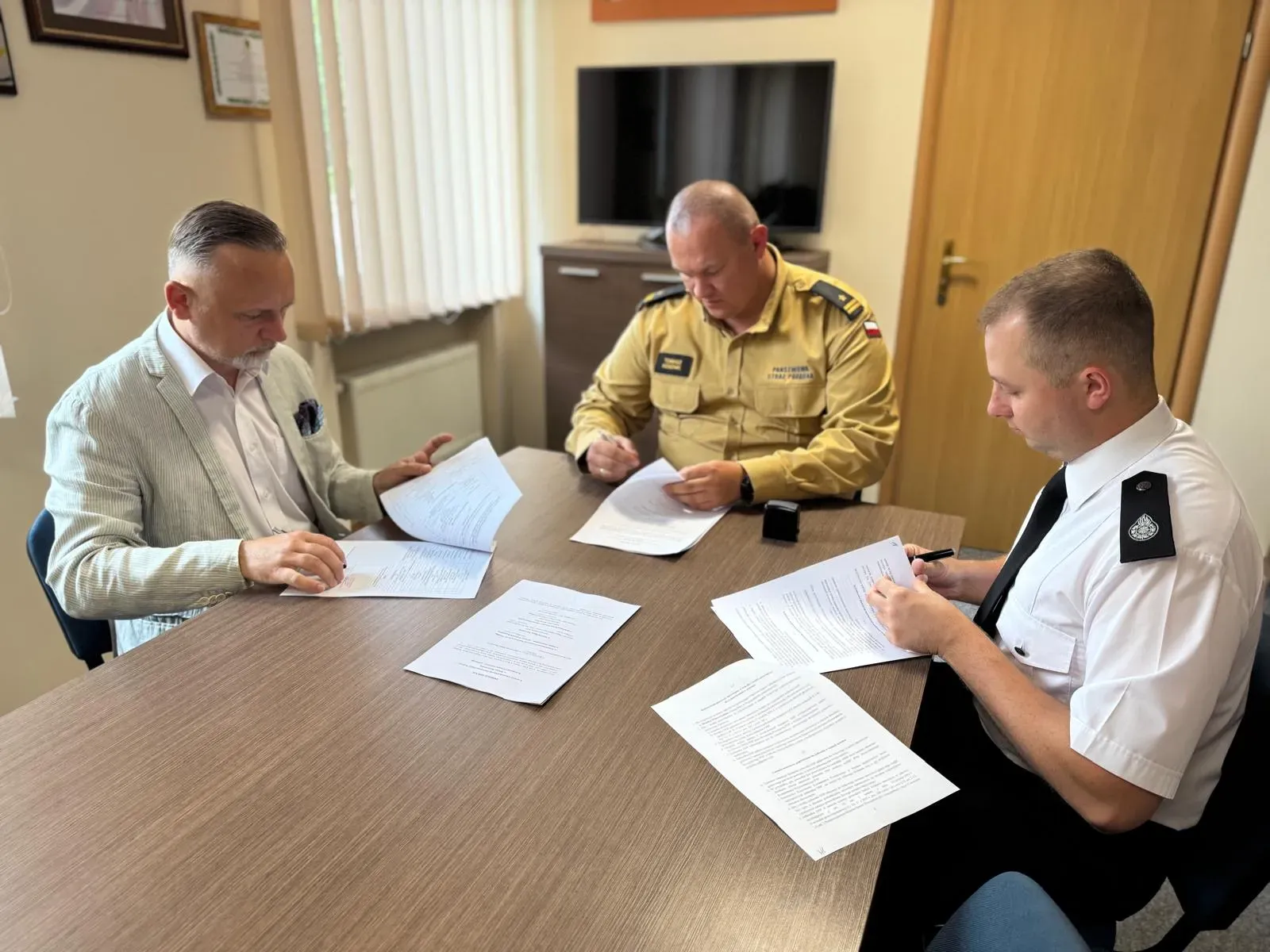 Oświetlone pomieszczenie. Trzech mężczyzn siedzi przy stole, przeglądając i podpisując dokumenty, z których jeden ma mundur służbowy straży pożarnej.
