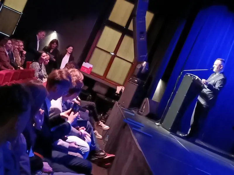 Mężczyzna przemawia z mównicy na scenie oświetlonej niebieskim światłem, a publiczność siedzi i słucha.