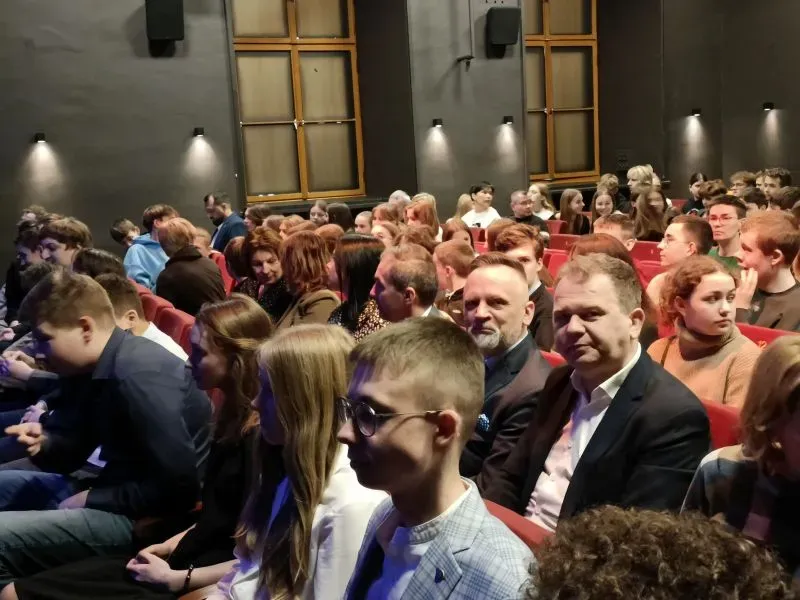 Grupa osób siedzących w rzędach czerwonych foteli w sali z szarymi ścianami i dużymi oknami.