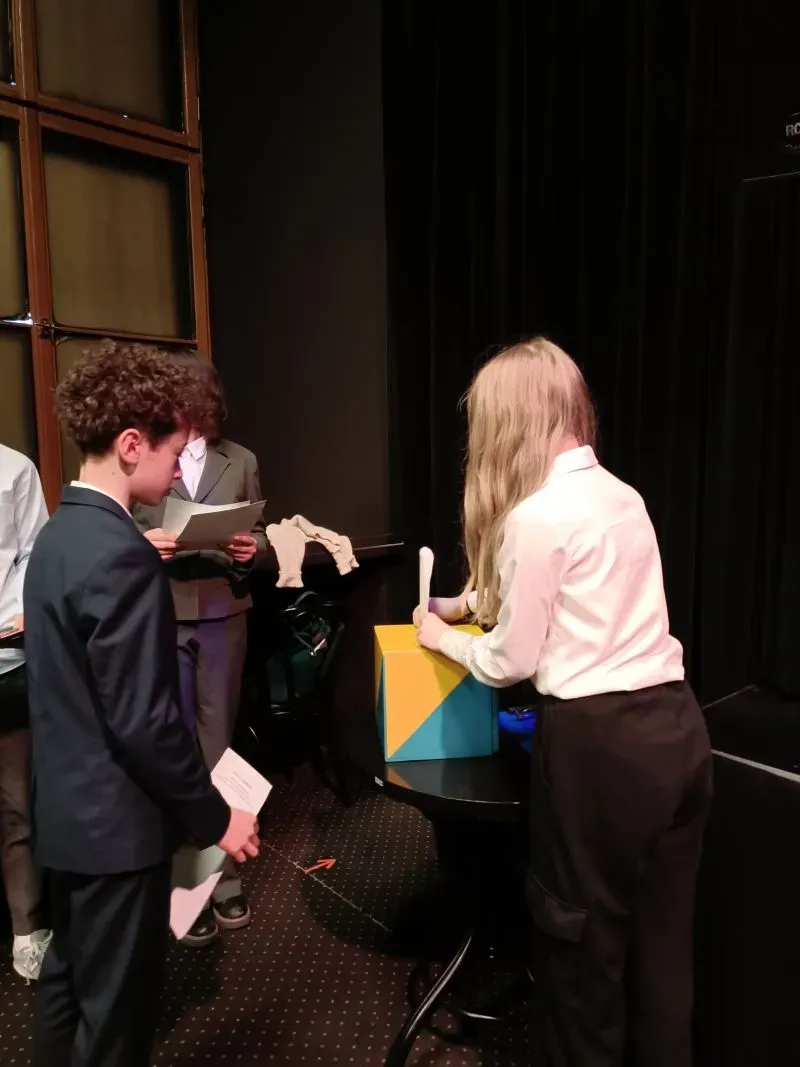 Dwóch nastolatków ubranych formalnie stoi obok stołu, na którym znajduje się kolorowa kostka, jedna osoba trzyma kartkę papieru.