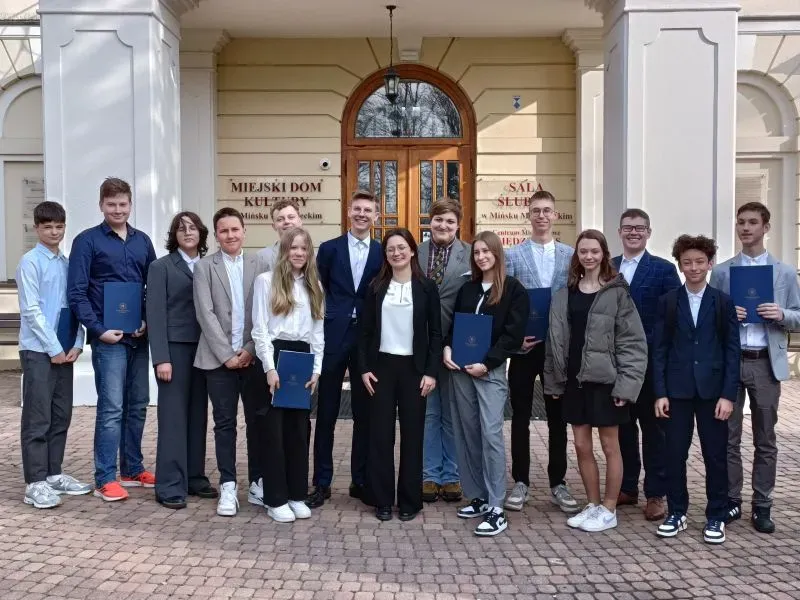 Grupa nastolatków i dorosłych ubranych formalnie stoi przed wejściem do budynku Miejskiego Domu Kultury.