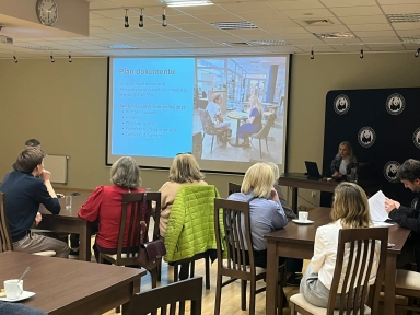 Osiem osób siedzi przy drewnianych stołach w jasnej sali konferencyjnej z rzutnikiem. Na środku ściany wisi ekran z niebieskim slajdem zawierającym listę punktów. Po prawej stronie przy stole siedzi prowadząca przy otwartym laptopie. Kilka kurtek jest zarzuconych na krzesła, na stolikach stoją filiżanki i dokumenty.