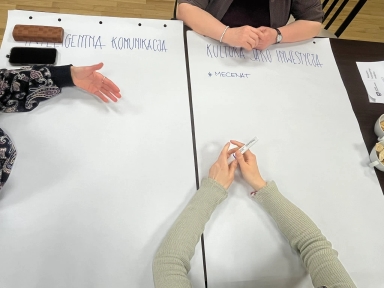 Dwie duże białe kartki rozłożone na ciemnym stole. Trzy pary rąk przy kartkach: jedna para po lewej gestykuluje, druga u góry ma złożone dłonie z bransoletką, trzecia u dołu trzyma marker. Na stole leży telefon i etui. Po prawej dwie filiżanki z przekąskami. U góry widoczna drewniana podłoga.