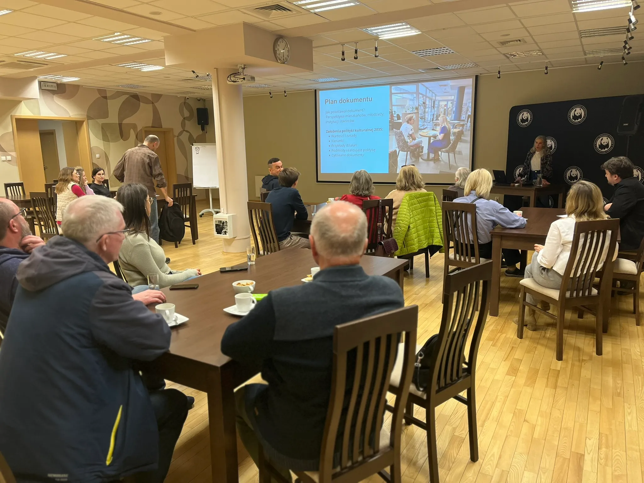 Kilkanaście osób siedzi przy drewnianych stołach w jasnej sali konferencyjnej. Na przodzie pokoju wyświetlany jest slajd na dużym ekranie, a kobieta stoi obok ekranu i prowadzi prezentację. Wnętrze ma drewnianą podłogę, jasne sufity z oświetleniem i beżowe ściany.