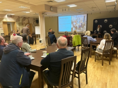 Kilkanaście osób siedzi przy drewnianych stołach w jasnej sali konferencyjnej. Na przodzie pokoju wyświetlany jest slajd na dużym ekranie, a kobieta stoi obok ekranu i prowadzi prezentację. Wnętrze ma drewnianą podłogę, jasne sufity z oświetleniem i beżowe ściany.