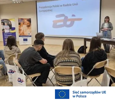 Grupa osób siedzi na krzesłach podczas prezentacji, na ekranie widoczny jest napis „Prezydencja Polski w Radzie Unii Europejskiej” oraz logo.