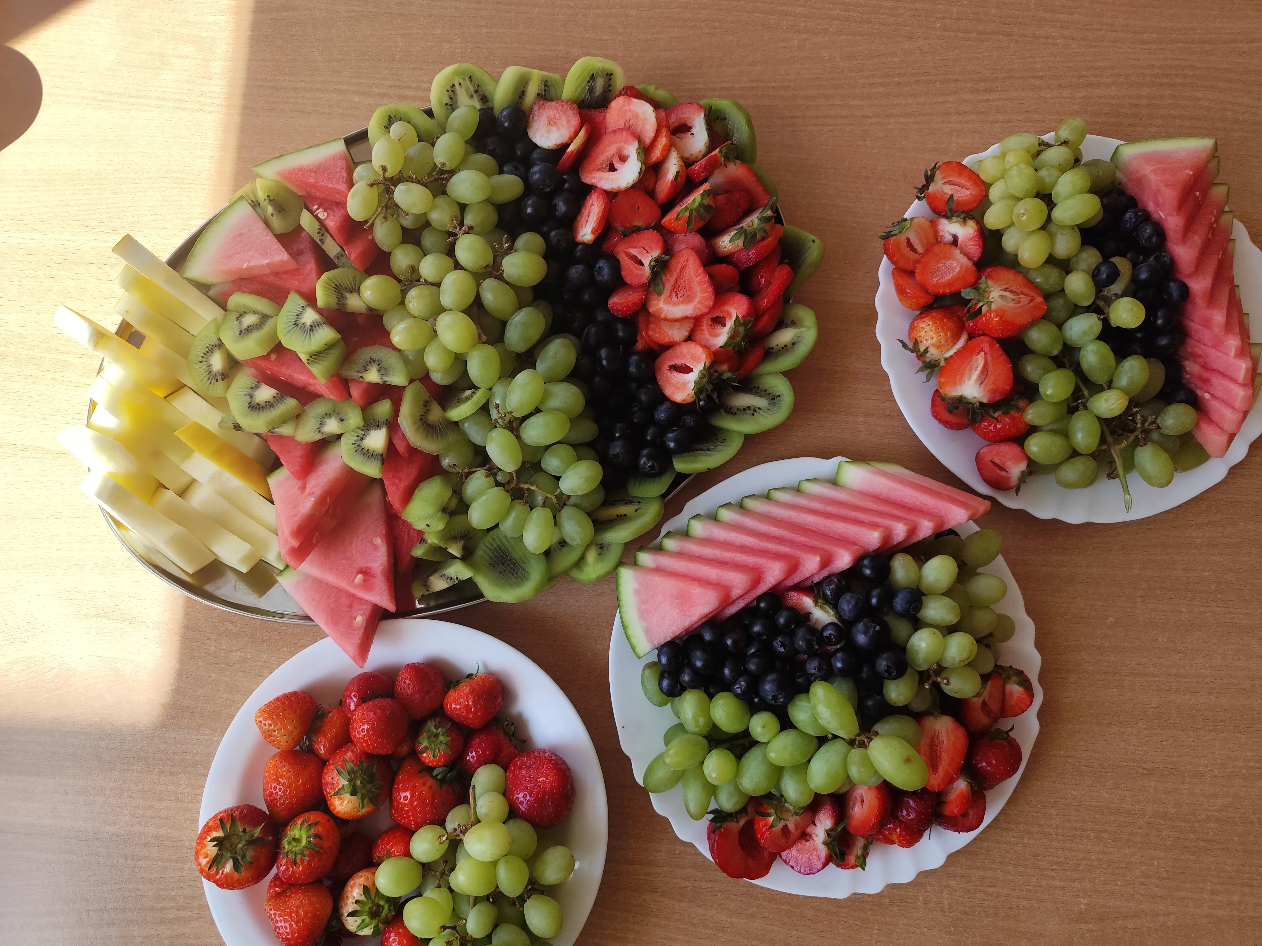 Pięć talerzy z owocami na jasnobrązowym, drewnianym stole w świetle dziennym. Na talerzach zielone winogrona, czerwone truskawki, ciemne jagody, trójkątne plasterki arbuza, plasterki kiwi i paski żółtego melona ułożone w porcje.