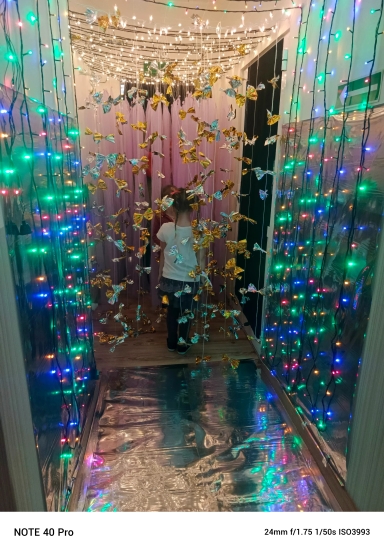 Dziecko stoi tyłem i przechodzi korytarzem ozdobionym wiszącymi złotymi i srebrnymi motylkami. Po bokach i nad głową wiszą kolorowe lampki choinkowe, a na podłodze i ścianach błyszcząca folia. W głębi wisi fioletowa zasłona, wnętrze jest oświetlone nocnym, dekoracyjnym światłem.