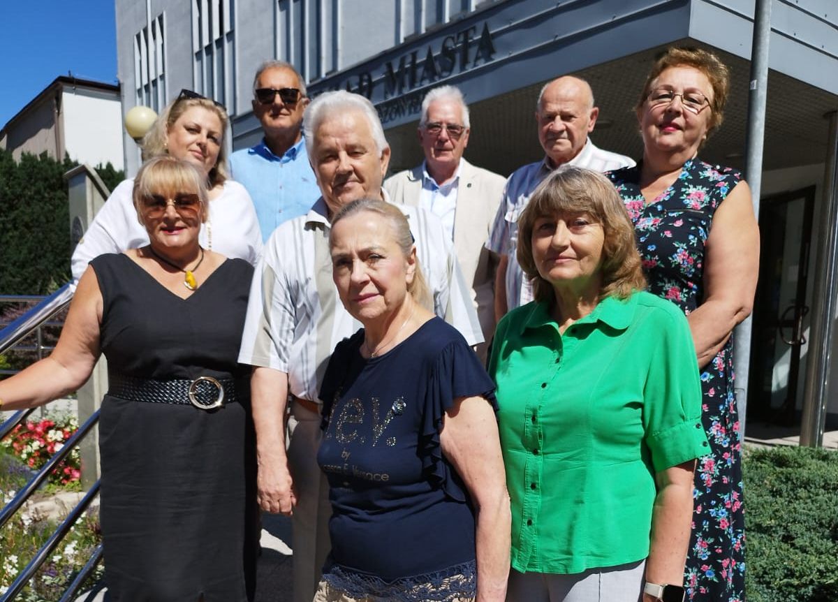 Na zdjęciu grupa dziesięciu osób stoi na zewnątrz, na tle budynku z napisem, w słoneczny dzień. Pięć osób znajduje się z przodu, a pięć z tyłu, w różnych kolorach ubrań.