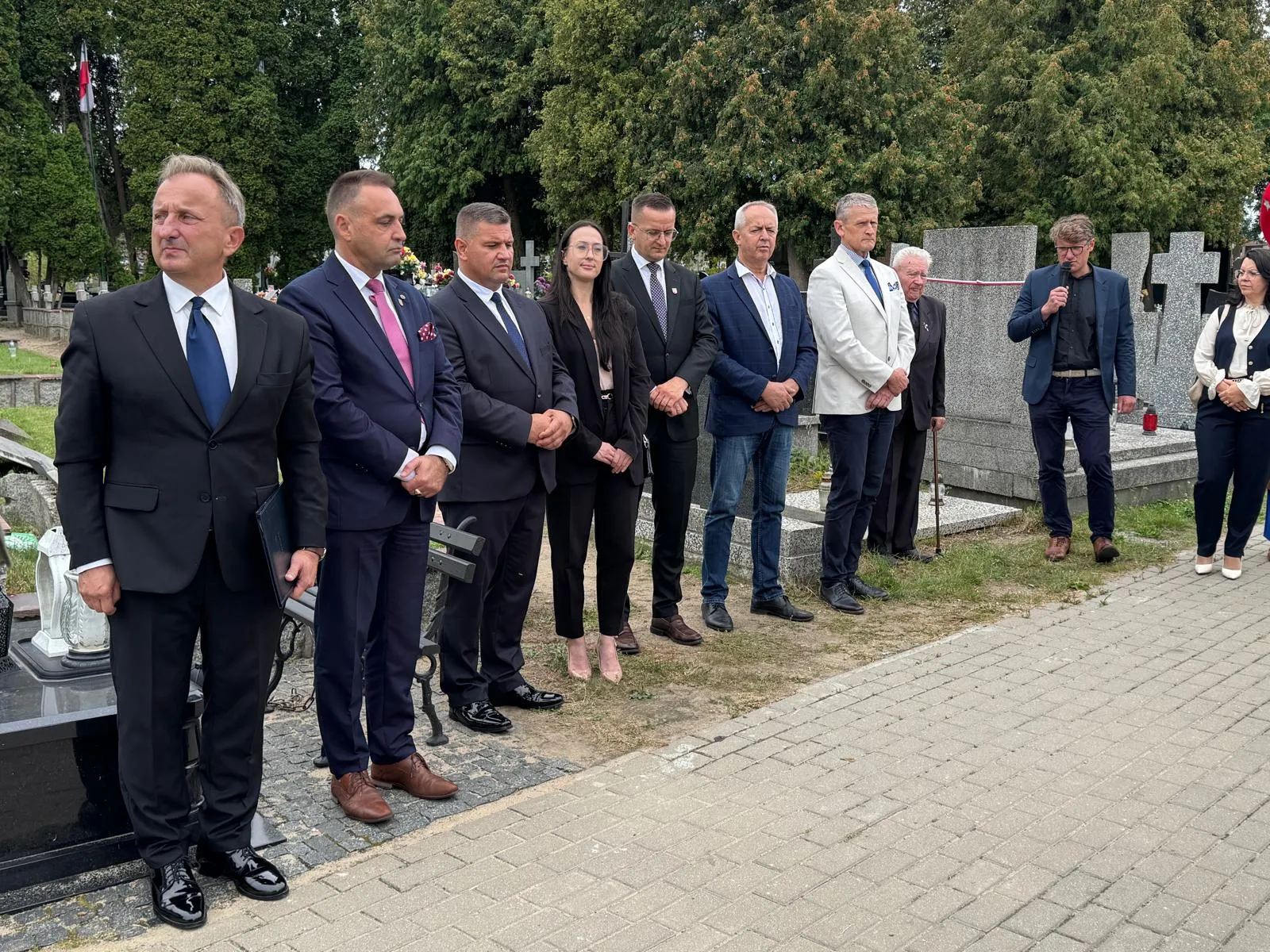 Dziesięć osób stoi na zewnątrz na cmentarzu przy pomnikach i nagrobkach, jedna osoba mówi do mikrofonu.