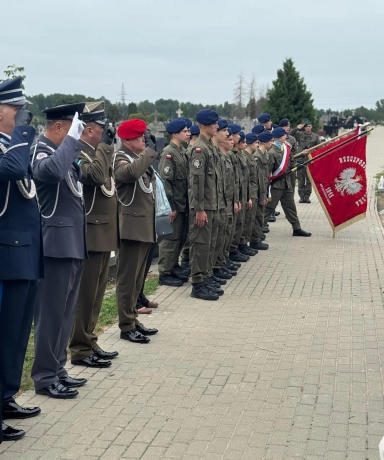 Żołnierze i oficerowie w mundurach ustawieni w szeregu na cmentarzu podczas uroczystego apelu, jeden z nich trzyma chorągiew RP.