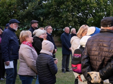 Grupa kilkunastu osób stoi na zewnątrz na trawie, ubranych w jesienne kurtki i czapki, w tle drzewa.