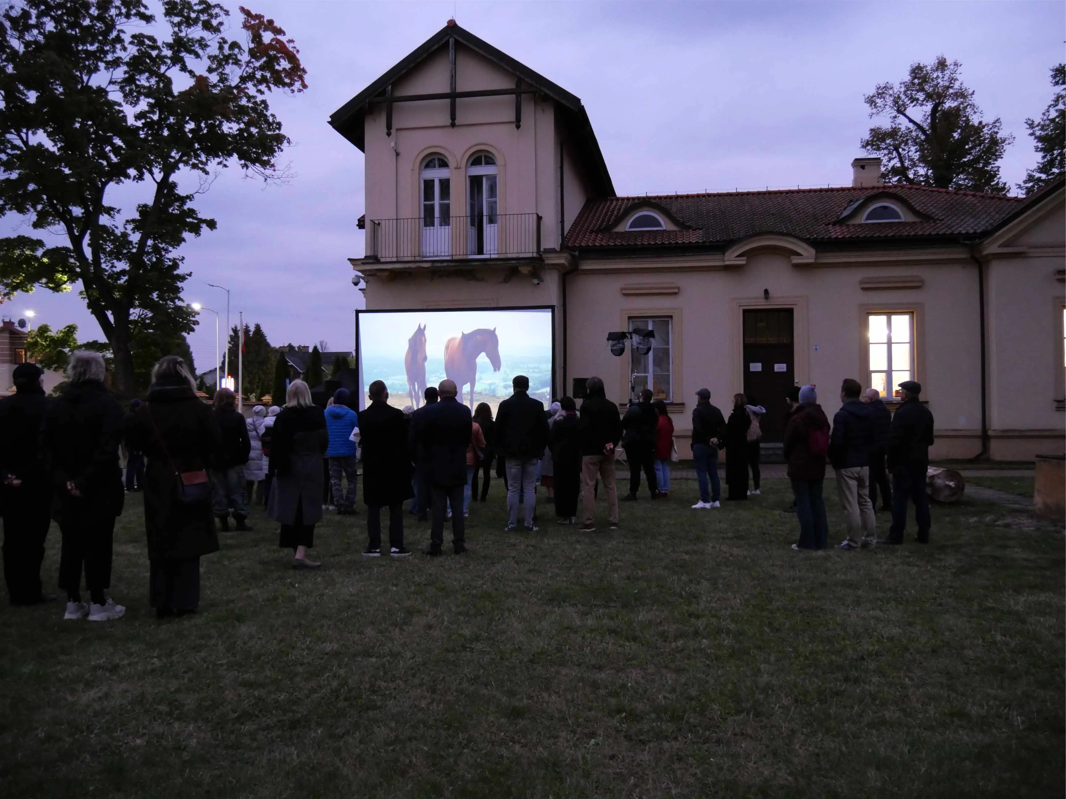 Grupa osób stoi na trawniku przed dużym budynkiem o jasnej elewacji podczas wieczoru. Przed nimi znajduje się ekran projekcyjny, na którym wyświetlane jest wideo z dwoma końmi. W tle widać drzewa.