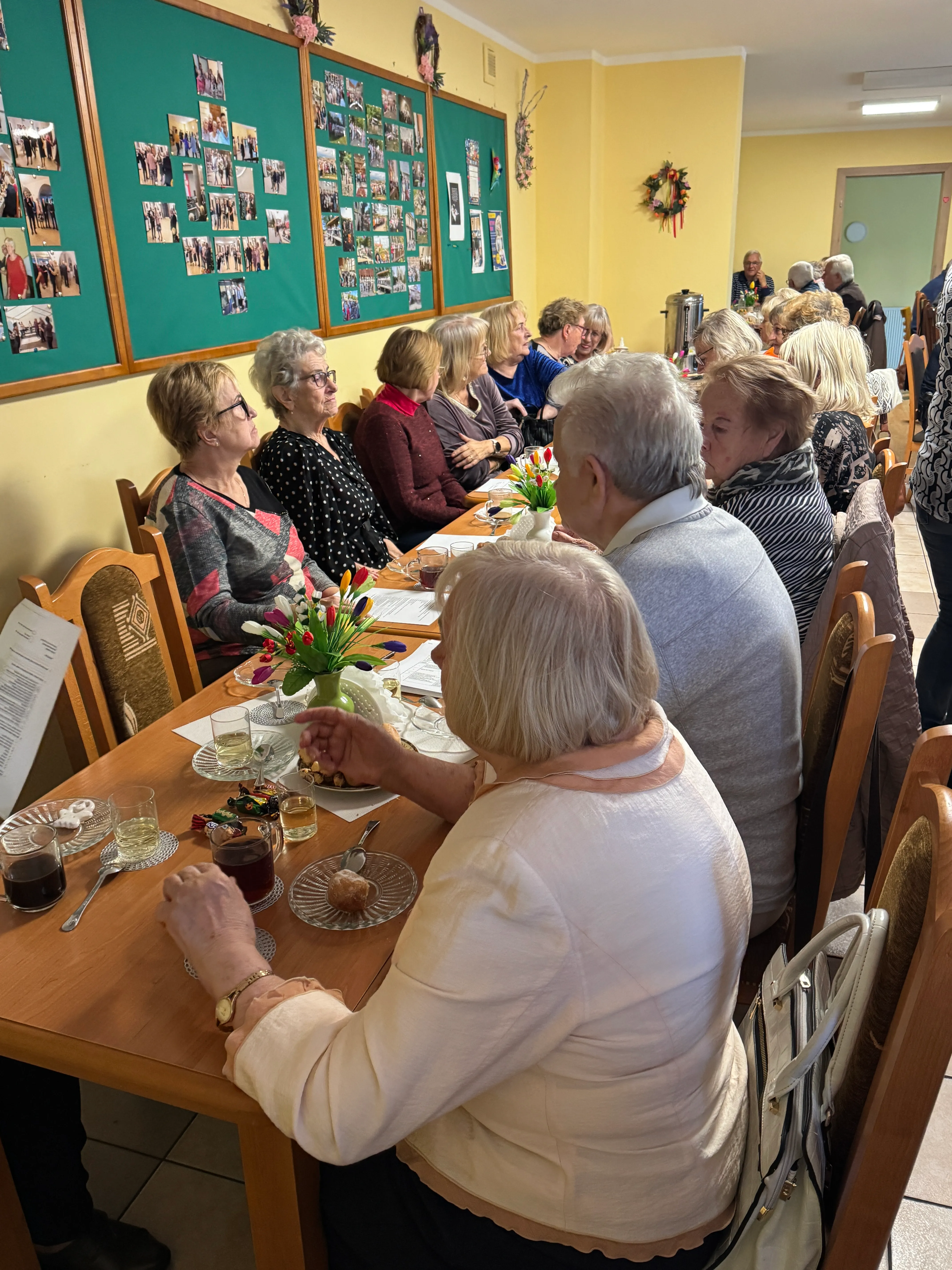 Grupa osób siedzi przy długim stole w jasnym pomieszczeniu. Na stole stoją naczynia z napojami, talerze z przekąskami i dekoracje z kolorowych kwiatów. Na ścianie wiszą tablice z licznymi zdjęciami. W tle widać drzwi i kolejne osoby siedzące przy stolikach.
