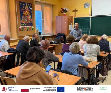 Grupa starszych osób siedzi przy ławkach w klasie, słuchając mężczyzny przemawiającego przy tablicy. W tle na ścianie wisi krzyż, szafa oraz dekoracje świąteczne.