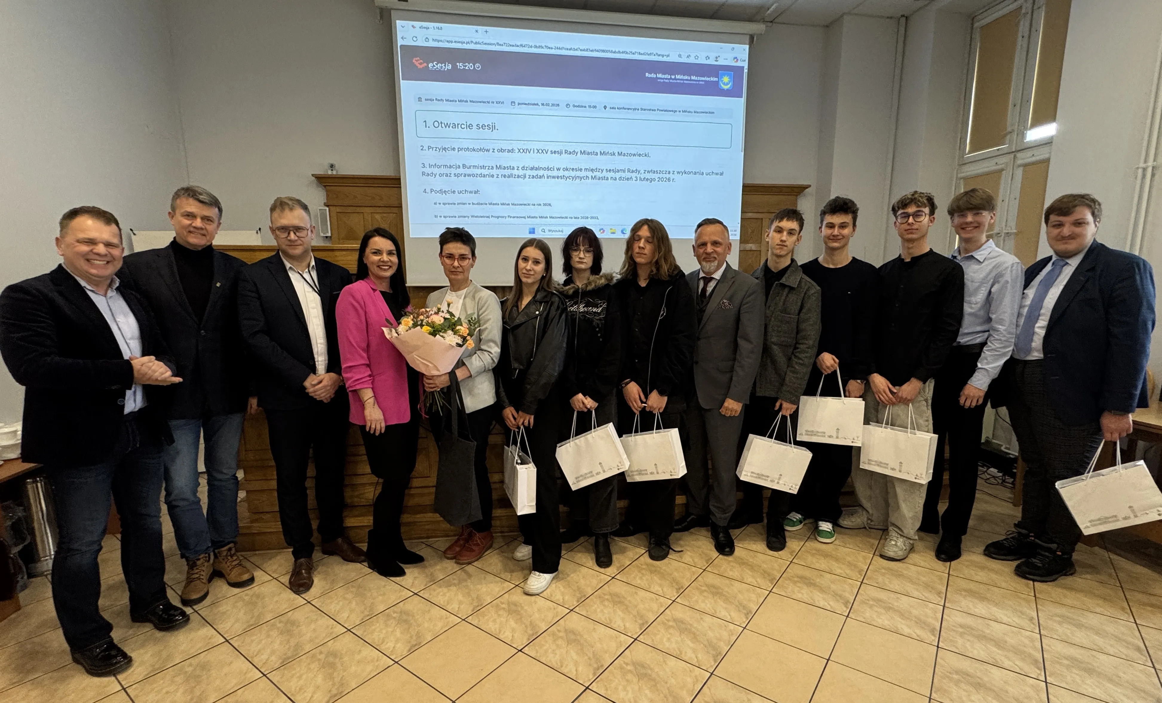 Piętnaście osób stoi w rzędzie w sali konferencyjnej przed dużym rzutnikiem. Kilka osób trzyma białe torby upominkowe, jedna osoba trzyma bukiet kwiatów. W tle jasne ściany, drewniany panel i tłumaczony slajd na ekranie.