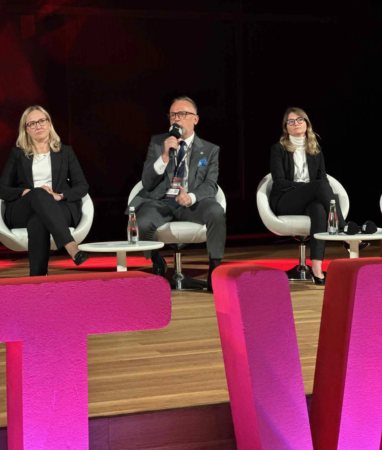 Trzy osoby siedzą na scenie na białych krzesłach z mikrofonem i butelkami wody na stołkach, mężczyzna po środku mówi, kobiety po bokach słuchają.