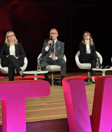 Trzy osoby siedzą na scenie na białych krzesłach z mikrofonem i butelkami wody na stołkach, mężczyzna po środku mówi, kobiety po bokach słuchają.