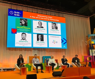 Pięć osób siedzi na krzesłach na scenie konferencyjnej z pomarańczową podłogą i jasną zasłoną w tle. Nad nimi duży ekran wyświetla prezentację z tytułem „Empowering Cities: A New all-in-one tool for Partnerships” oraz zdjęciami i nazwiskami prelegentów. Scena jest oświetlona ciepłym światłem.