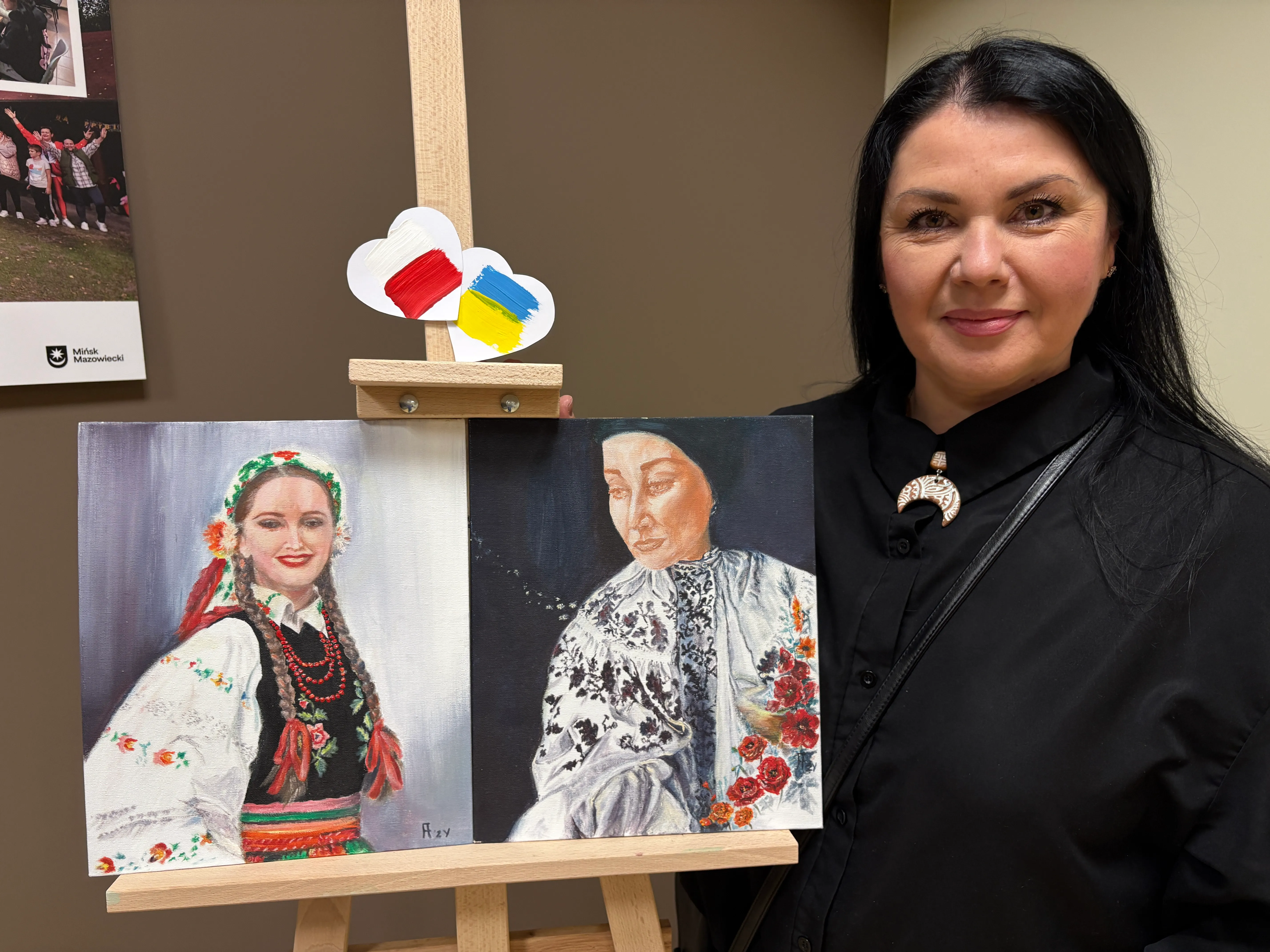 Kobieta stoi obok sztalugi w pomieszczeniu wewnętrznym, ma czarne włosy i czarną bluzkę. Na sztaludze umieszczony jest dwuczęściowy obraz: po lewej młoda kobieta w kolorowym stroju ludowym z warkoczami i koralami, po prawej portret starszej kobiety w haftowanej, jasnej bluzce z czerwonymi kwiatami. U góry sztalugi przybite są dwa papierowe serca z namalowanymi flagami Polski (czerwono-biała) i Ukrainy (niebiesko-żółta).