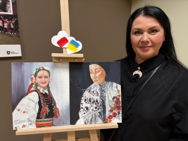 Kobieta stoi obok sztalugi w pomieszczeniu wewnętrznym, ma czarne włosy i czarną bluzkę. Na sztaludze umieszczony jest dwuczęściowy obraz: po lewej młoda kobieta w kolorowym stroju ludowym z warkoczami i koralami, po prawej portret starszej kobiety w haftowanej, jasnej bluzce z czerwonymi kwiatami. U góry sztalugi przybite są dwa papierowe serca z namalowanymi flagami Polski (czerwono-biała) i Ukrainy (niebiesko-żółta).