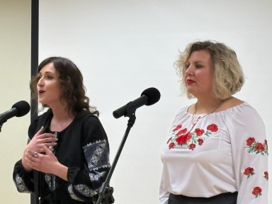 Dwie kobiety stoją obok siebie przed mikrofonami. Lewa kobieta ma ciemne kręcone włosy i czarną bluzkę z haftowanym wzorem, śpiewa i gestykuluje dłońmi. Prawa kobieta ma krótkie blond włosy i białą bluzkę z czerwonymi haftami, stoi przy mikrofonie. Jasne, gładkie tło wewnątrz pomieszczenia.